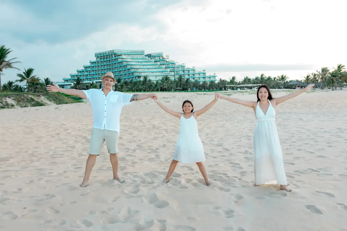 A joyful family holding hands on a sandy beach in Hội An, with a resort and palm trees in the background.